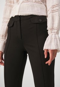 Pantalon noir cintré avec poches plaquées à rabats et boutons, porté avec un haut blanc en dentelle à manches longues avec poignets volantés.