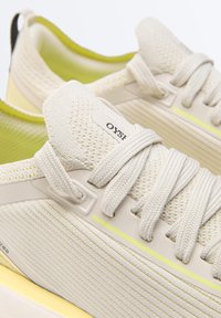 Gros plan sur des baskets beiges en tricot avec des lacets épais, des accents jaunes et des semelles rembourrées, mettant en valeur la texture et le détail de la marque sur la languette.