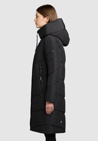 Frau in einem langen schwarzen, gepolsterten Mantel mit Kapuze, hohem Kragen und mit Reißverschlüssen versehenen Taschen, die im Profil vor einem schlichten, hellen Hintergrund steht.