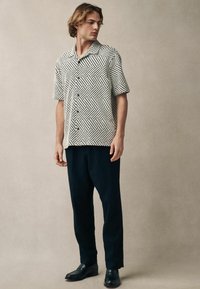 Camisa de manga corta con un patrón geométrico en blanco y negro, con botones en la parte delantera y un corte relajado, combinada con pantalones navy y mocasines negros.