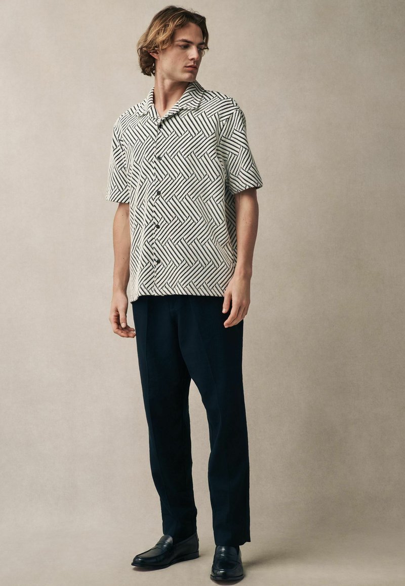 Camisa de manga corta con un patrón geométrico en blanco y negro, con botones en la parte delantera y un corte relajado, combinada con pantalones navy y mocasines negros.