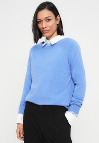 Femme aux cheveux châtains mi-longs portant un pull bleu clair par-dessus une chemise blanche à col et un pantalon noir, debout devant un fond blanc.