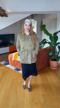 Νεαρή γυναίκα με μακριά μαλλιά, ντυμένη με μπεζ καμπαρντίνα, μαύρη φούστα και γκρι παπούτσια, στέκεται σε ένα σαλόνι με πορτοκαλί καναπέ και μεγάλο φυτεμένο φυτό.