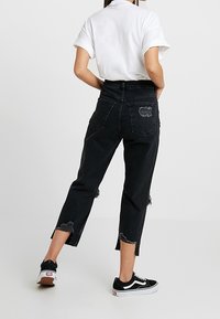 Jeans neri a vita alta, tagliati e con orli sfrangiati, dotati di una tasca posteriore con logo. Abbinati a una camicia bianca e sneakers nere.