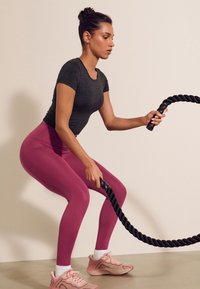 Sportliches Outfit mit einem eng anliegenden dunkelgrauen T-Shirt und pinken Leggings. Die Person hält ein schwarzes Battle Rope, trägt hellrosa Turnschuhe mit weißen Socken.