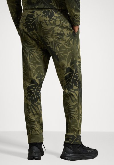 Zelené joggers s tropickým listovým vzorem, vyrobené z měkké látky, s postranními kapsami a přiléhavými manžetami. Zobrazené černé tenisky.