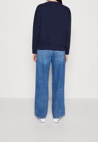 Sudadera azul marino con puños de canalé, combinada con pantalones vaqueros de pierna ancha de acabado desgastado y dos bolsillos traseros. Se llevan zapatillas blancas.
