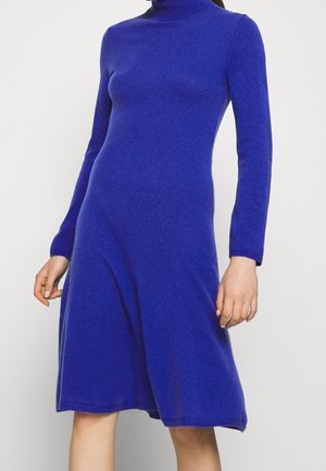 Mujer con un vestido suéter azul ajustado de manga larga, hasta la rodilla y con cuello alto, sobre un fondo blanco liso.