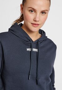 Mörkblå hoodie med känguruficka, dragsko i huvan och en vit tryckt logotyp "HUMMEL" på bröstet. Tillverkad av mjukt tyg.