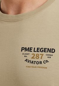 Beige bavlněné tričko s kulatým výstřihem. Obsahuje černý a zlatý tisk s textem: "PME LEGEND," "FLIGHT NO. 287," "AVIATOR CO.," "FIND YOUR FREEDOM."