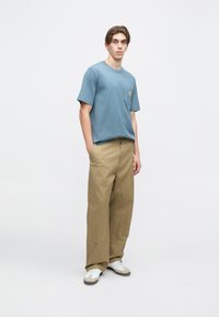 Světle modré bavlněné tričko s kapsou na prsou, spárované s pohodlnými béžovými kalhotami. Oblečení zahrnuje bílé tenisky s hnědými akcenty.