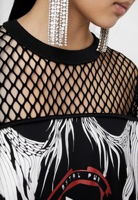 Gros plan d'une personne portant de longues boucles d'oreilles en cristal et un haut en résille noire avec un imprimé graphique noir et blanc en dessous.