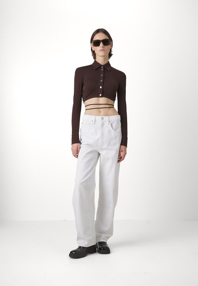 Barna, cropped, hosszú ujjú felső gombokkal, magas derékú fehér farmernadrág és fekete slip-on cipő, semleges háttér előtt.