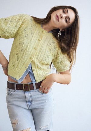 Femme aux longs cheveux bruns portant un cardigan jaune tricoté à manches courtes, un jean bleu clair déchiré et des boucles d'oreilles argentées, posant avec la main sur la hanche.