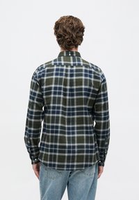 Göndör hajú férfi zöld, tengerész kék és szürke kockás ingben és kék farmernadrágban áll, háttérben fehér fal.