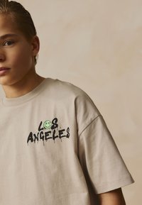 Beiges Baumwoll-T-Shirt mit schwarzem Graffiti-Print "LOS ANGELES" und einem kleinen grünen Smiley. Lockerer Schnitt und kurze Ärmel.