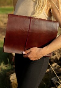 Hnědá kožená kabelka ve stylu obálky držena v ruce, s hladkým povrchem a kontrastním vínovým elastickým uzávěrem, s minimalistickým designem.