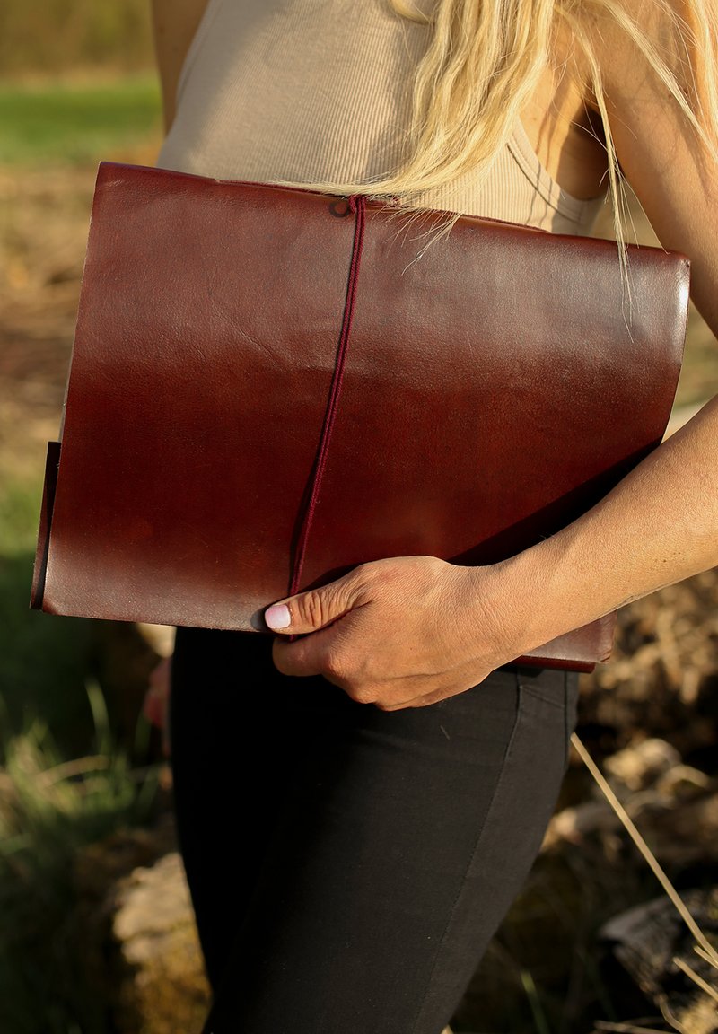 Hnědá kožená kabelka ve stylu obálky držena v ruce, s hladkým povrchem a kontrastním vínovým elastickým uzávěrem, s minimalistickým designem.