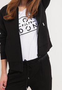 Sort zip-jakke med glat stof, rund halsudskæring og lange ærmer, lagd over en hvid t-shirt med fed grafisk tekst.