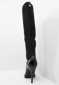 Bota negra sobre la rodilla con una textura lisa y elegante y un tacón de charol brillante. Presenta una costura vertical y un sutil detalle metálico.