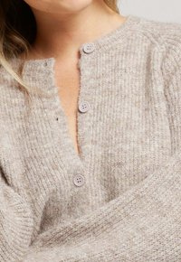 Cardigan beige clair en tricot texturé doux, avec un col en V, cinq boutons lilas et des poignets côtelés.