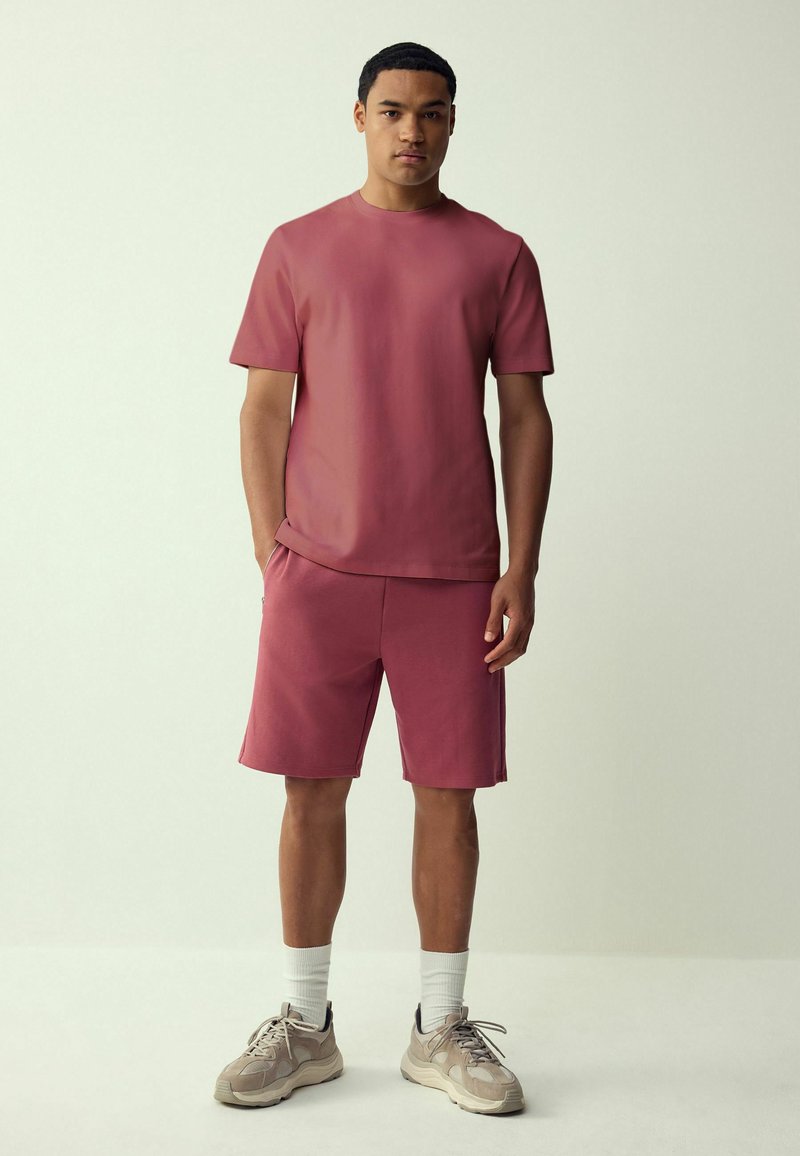 Un giovane uomo in piedi di fronte, indossa una maglietta a maniche corte e pantaloncini abbinati color rosa polvere, con calzini bianchi e scarpe da ginnastica beige.