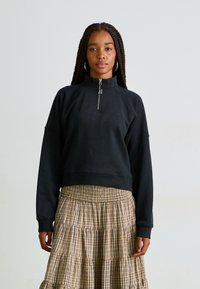 Sudadera negra con media cremallera y corte relajado, combinada con una falda de cuadros beige en capas con cintura elástica.