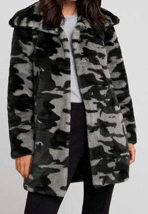 Vrouw draagt een zwart-grijze camouflage bontjas van gemiddelde lengte over een lichtgrijze top en zwarte broek, staand tegen een effen achtergrond.