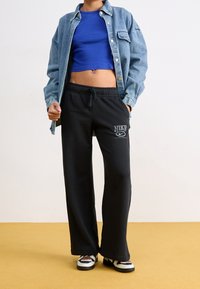 Blauwe cropped top, een denim overhemd met lange mouwen en zwarte sweatpants met het Nike-logo. De sweatpants hebben een losse pasvorm, en de schoenen hebben een speelse uitstraling.