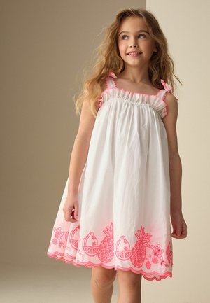 Junges Mädchen mit langen blonden Haaren, das ein weißes ärmelloses Kleid mit rosa Fruchtmustern und Rüschensaum trägt, lächelt und blickt zur Seite.