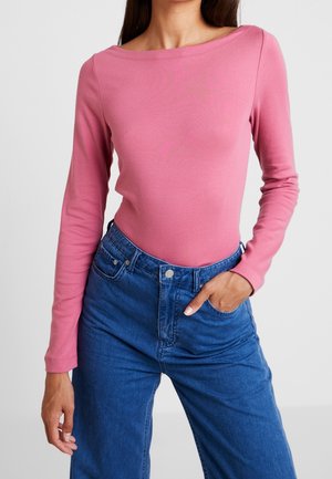 Mujer con una camiseta rosa ajustada de manga larga y vaqueros azules de tiro alto con una mano en el bolsillo sobre un fondo liso.