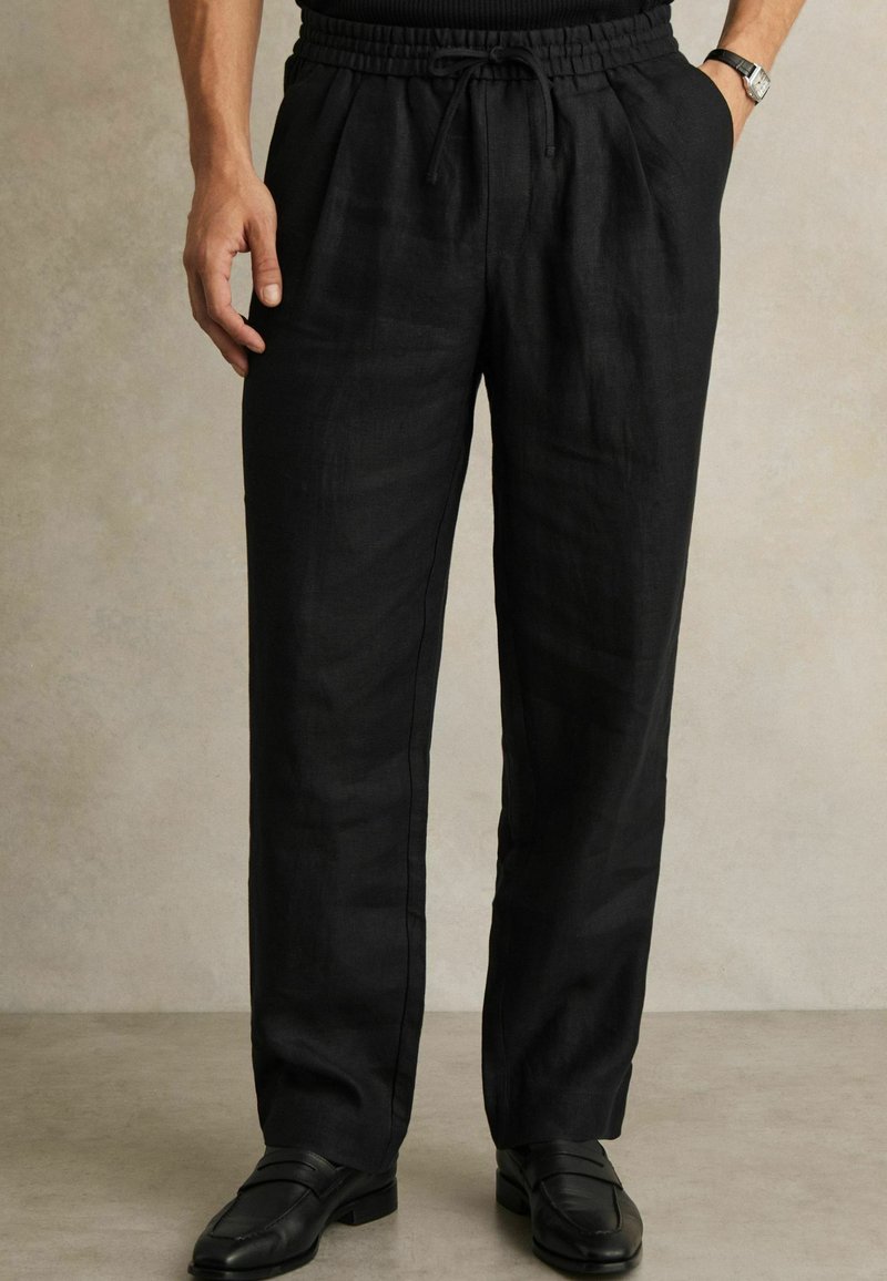 Fekete lenvászon nadrág, elasztikus derékpánttal és húzózsinórral, oldalsó zsebekkel és laza szabással, fekete elegáns cipőkkel párosítva.