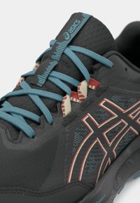 Zapatilla deportiva gris con parte superior texturizada, cordones azules y acentos rojos. Presenta un logotipo prominente y detalles en patrón en el lateral.