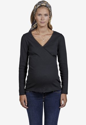 Schwarzes Langarm-Wickelshirt mit Spitzenbesatz am Ausschnitt und an den Ärmeln, kombiniert mit dunkelblauen Jeans. Das Shirt ist für die Schwangerschaftsbekleidung entworfen.