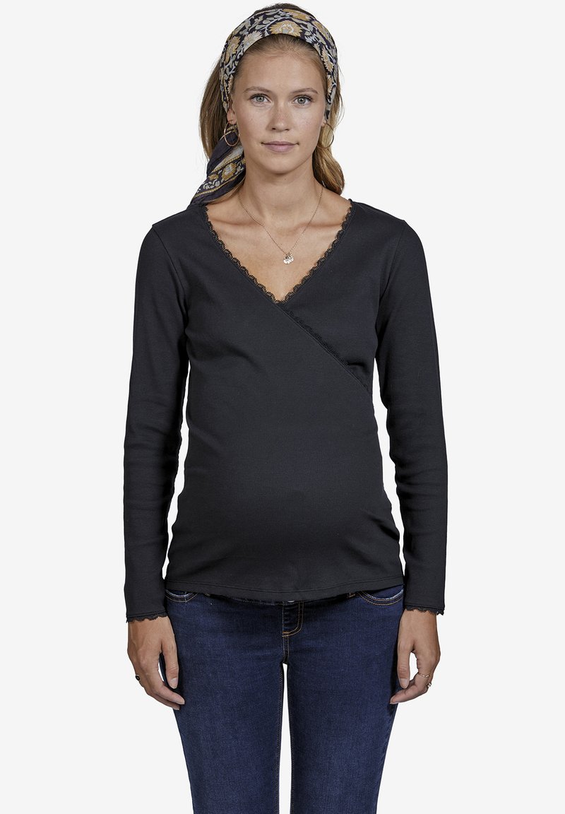 Schwarzes Langarm-Wickelshirt mit Spitzenbesatz am Ausschnitt und an den Ärmeln, kombiniert mit dunkelblauen Jeans. Das Shirt ist für die Schwangerschaftsbekleidung entworfen.