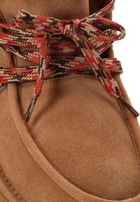 Bota de ante marrón con punta redondeada y complejas cintas trenzadas multicolores atadas en un lazo, que presenta una textura suave y costuras reforzadas.