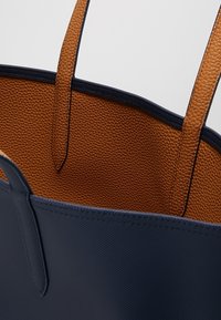 Sac fourre-tout bleu marine avec intérieur en cuir marron texturé et poignées marron assorties, présentant des coutures détaillées le long des bords.