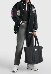 Chaqueta varsity negra con parches bordados, vaqueros rectos grises, una bolsa tote negra y zapatillas blancas con acentos de colores.