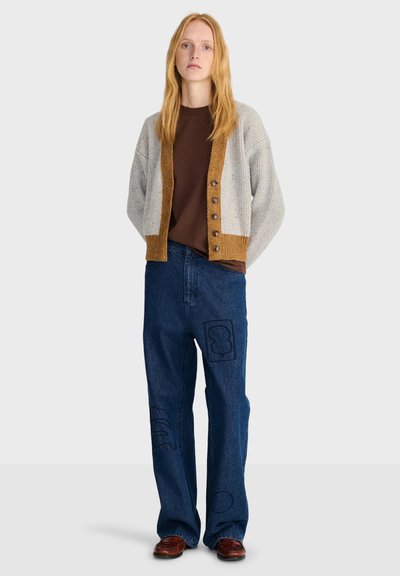 Femme aux longs cheveux blonds portant un cardigan gris et marron boutonné, une chemise marron, un jean bleu avec des patchs et des chaussures marron, debout les mains dans le dos.