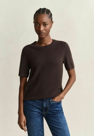 Frau mit geflochtenem Haar, die einen dunkelbraunen Pullover mit kurzen Ärmeln und blaue Jeans trägt, steht mit einer Hand in der Tasche vor einem schlichten Hintergrund.