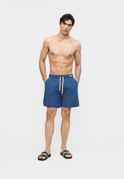 Mężczyzna bez koszulki, z rękami w kieszeniach, ubrany w niebieskie kąpielówki z żółtymi sznurkami i czarne klapki Moschino, stojący na białym tle.