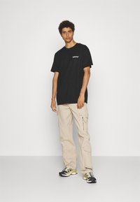Sort T-shirt med korte ærmer og hvidt logo på brystet; parret med lysebeige cargo-bukser med sidelommer og lynlåsdetailler.