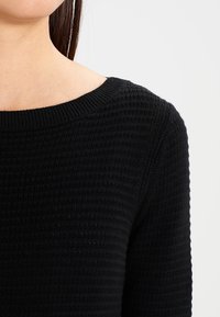 Svart stickad tröja med ribbad textur, bred rund halsringning och långa ärmar. Materialet verkar mjukt och bekvämt.