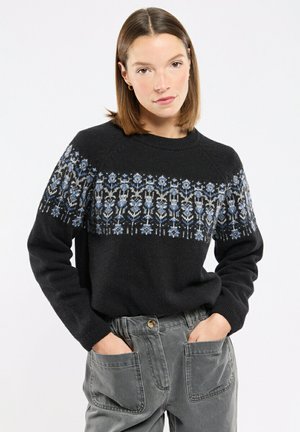 Donna che indossa un maglione nero con motivi floreali blu sul petto e pantaloni grigi a vita alta, in piedi con le mani nelle tasche.