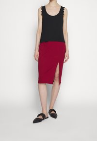 Femme porte un haut noir sans manches à bretelles festonnées, une jupe rouge longueur genou avec fente devant, et des ballerines noires à bout pointu.