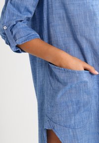 Light blue, linen shirt with roll-up sleeves, featuring a pocket and a curved hem. Texture appears slightly wrinkled, adding visual interest.