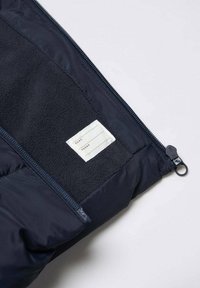 Oöppnad svart jacka som visar en mjuk innermembran med en sydd vit etikett för namn och telefoninformation.