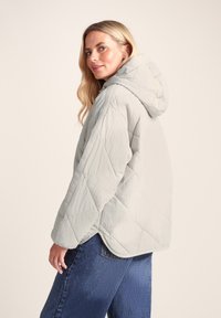 Quilted light grey hooded jacket, showcasing a relaxed fit, textured fabric, and slanted hem. Paired with dark blue denim jeans.