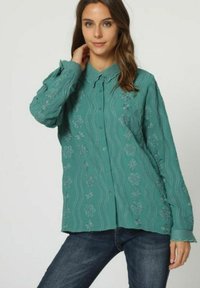 Camisa de botones en color azul verdoso con bordado floral y patrón ondulado; mangas largas con puños abotonados; corte relajado; textura suave.