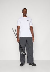 Uomo in t-shirt bianca Terrex e pantaloni grigi che tiene due bastoni da trekking neri contro uno sfondo chiaro uniforme.